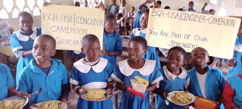 Two girls in school uniforms holding food plates and smiling in a classroom. Two girls in school uniforms holding food plates and smiling in a classroom.