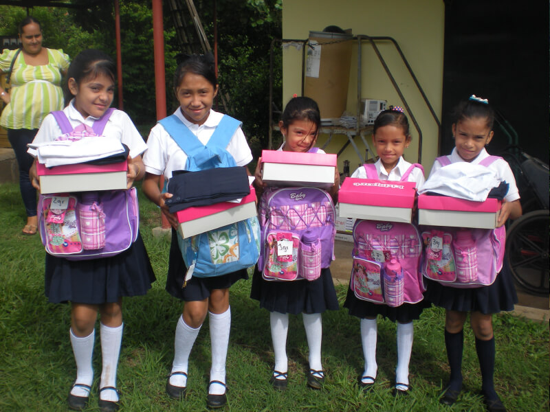 Five young girls in school uniforms holding school supply boxes outdoors.
