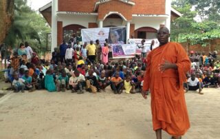 A monk stands in front of a large group gathered outside a building.