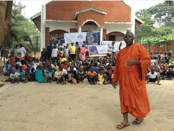 A monk stands in front of a large group gathered outside a building.