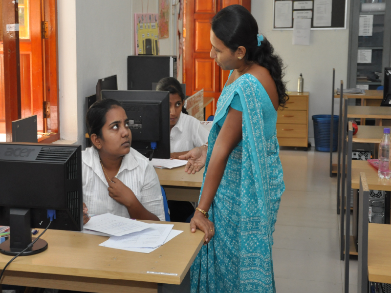 A woman in a blue saree talking to a seated woman in an office.