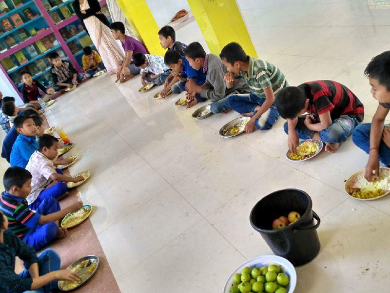 Children sitting in a row eating food from plates on the floor.