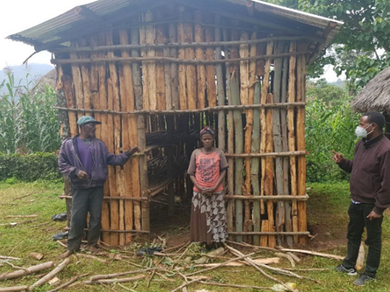 Two people standing in front of a small wooden structure in a rural setting.
