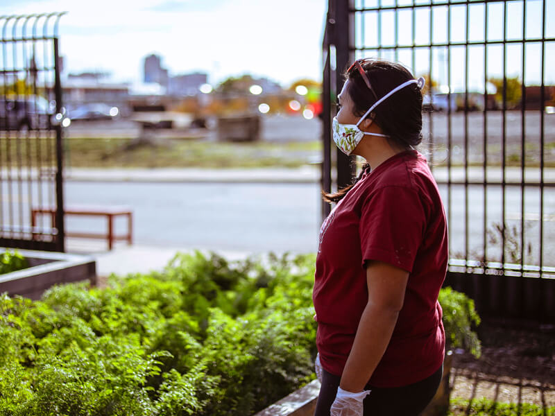 Person in red shirt wearing a mask and gloves near greenery and water.