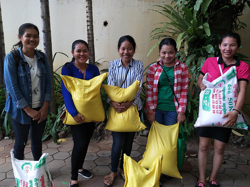 Four women happily holding large yellow bags, standing outdoors with greenery behind them.