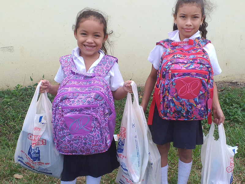 Two smiling schoolgirls holding bags and wearing backpacks.