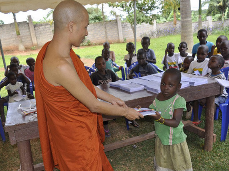 A monk hands a book to a young girl in an outdoor setting with children watching.