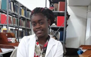 Young woman in a white lab coat smiling in a library setting.