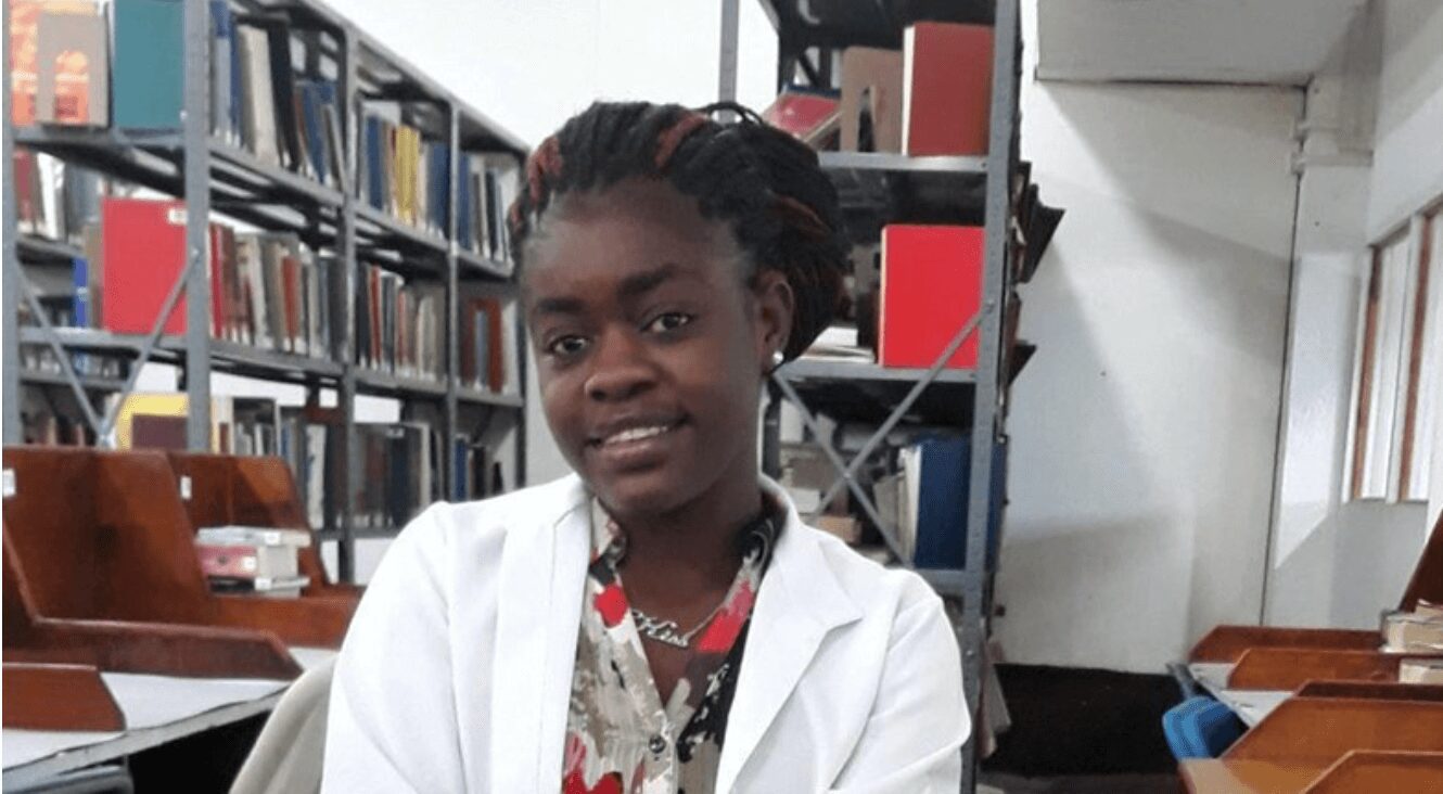 Young woman in a white lab coat smiling in a library setting.