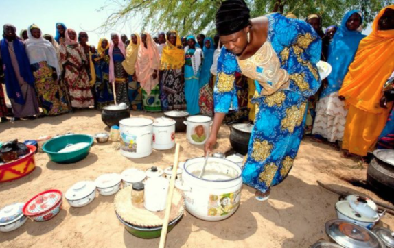 A woman stirring food in a large pot outdoors with people gathered around.