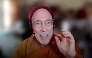 Elderly man speaking with expressive hand gesture and a blurred background.