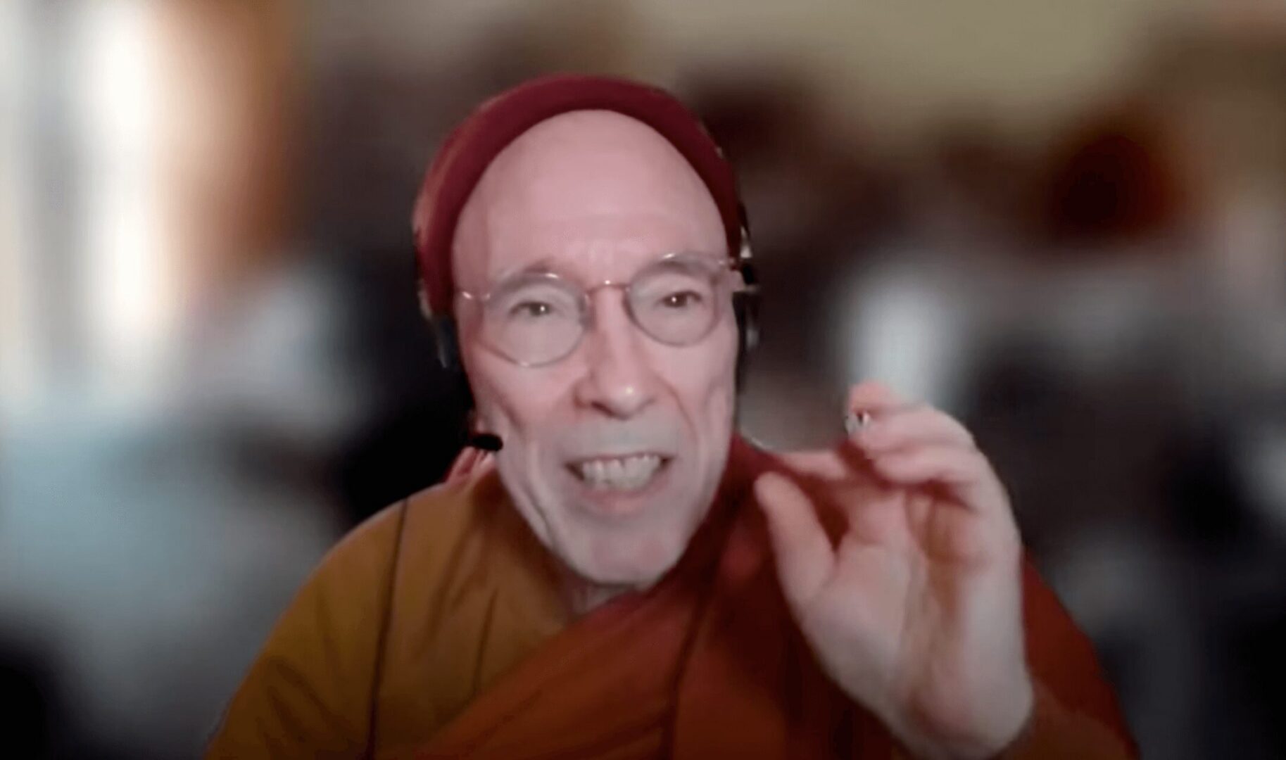 Elderly man speaking with expressive hand gesture and a blurred background.