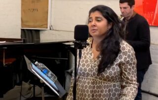 Woman singing in a music rehearsal room with a pianist in the background.