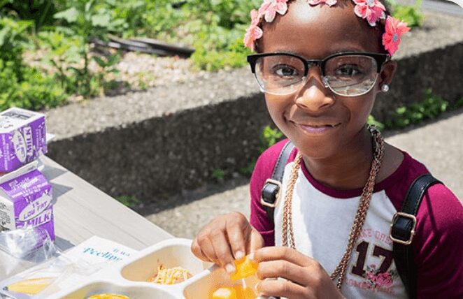 A girl with glasses and a flower headband enjoying a meal outdoors.