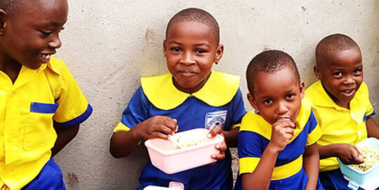 Four children in blue and yellow uniforms smiling with lunchboxes.