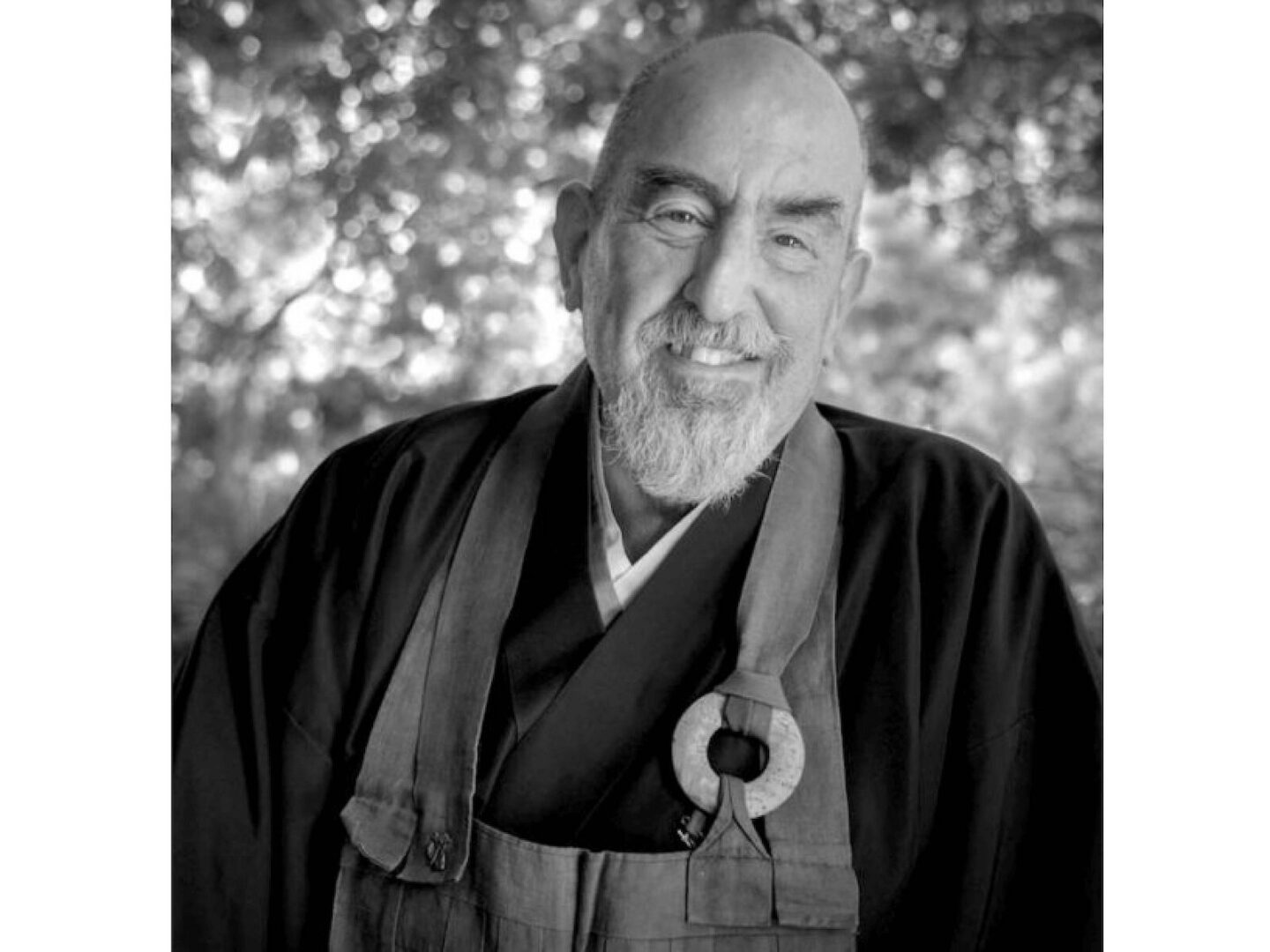A serene elderly man in traditional robes smiles warmly outdoors.